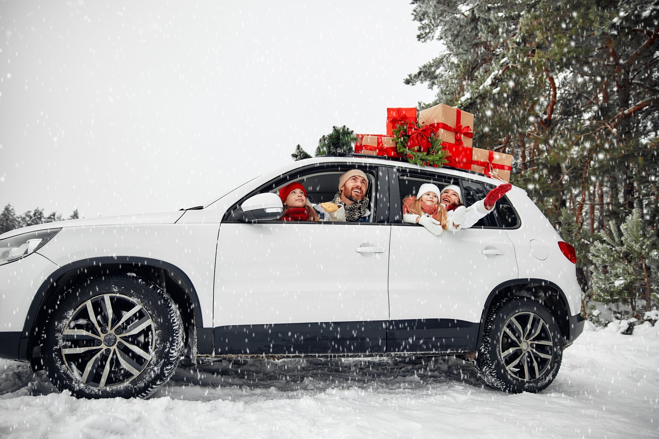 Happy family with children sitting in a car loaded with boxes of gifts and a Christmas tree against the background of a forest in snowy weather.